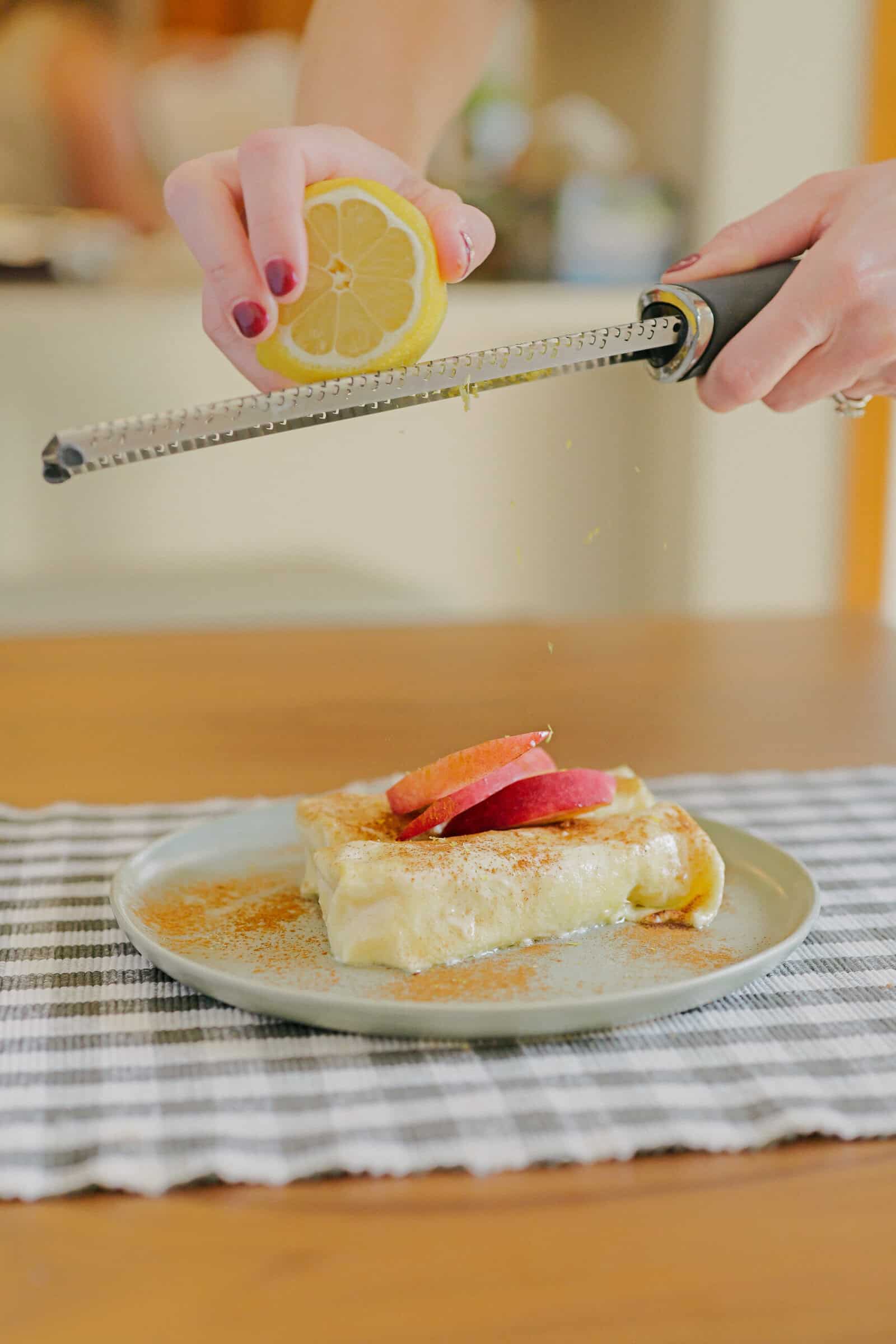 A person zesting a lemon over a plated dessert topped with sliced apples. The plate sits on a black-and-white checkered cloth on a wooden table.