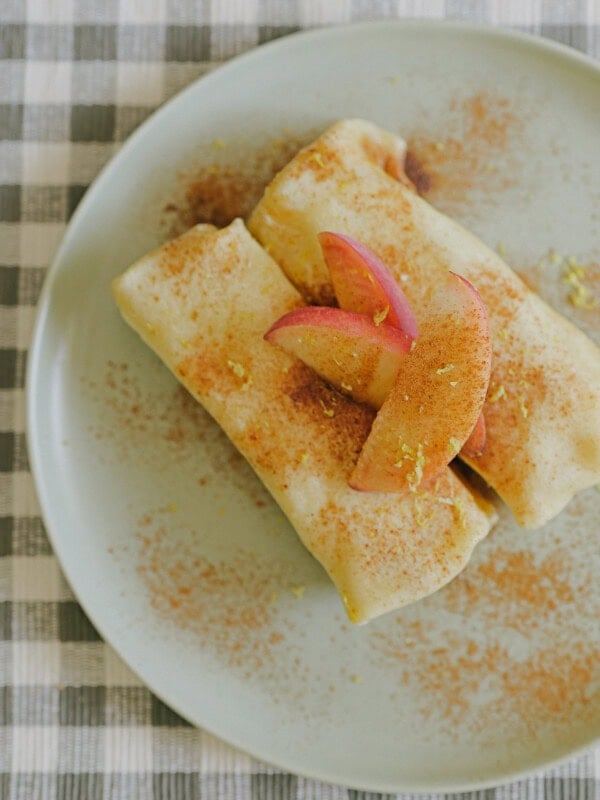 Two rolled crepes dusted with cinnamon on a pale plate, topped with three apple slices. Plate sits on a checkered tablecloth.