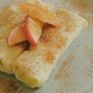 Two rolled crepes topped with apple slices, dusted with cinnamon and lemon zest, served on a light green plate on a checkered tablecloth.
