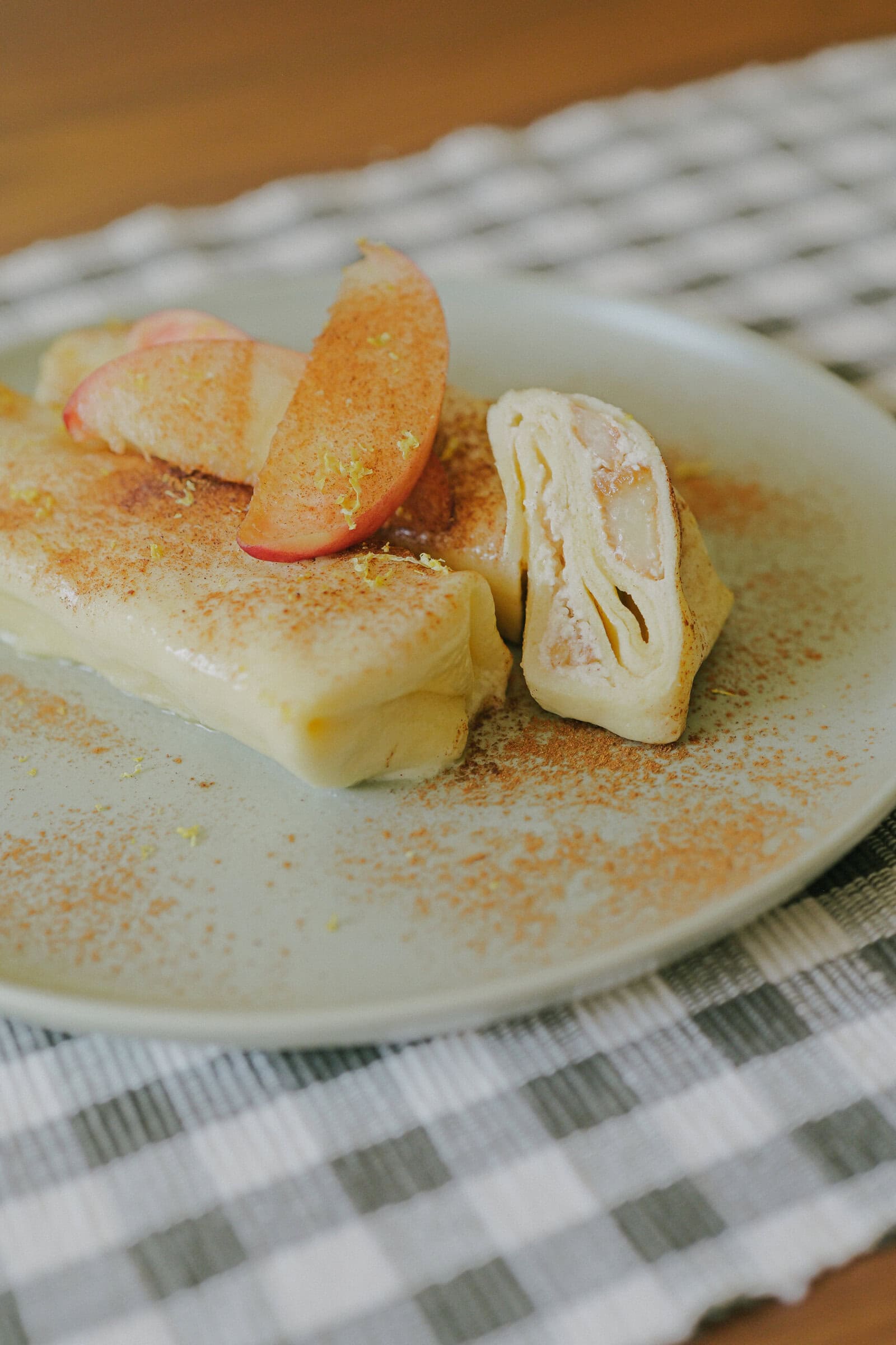 Two rolled crepes filled with sliced apples, topped with additional apple slices and sprinkled with cinnamon, served on a light green plate atop a checkered cloth.