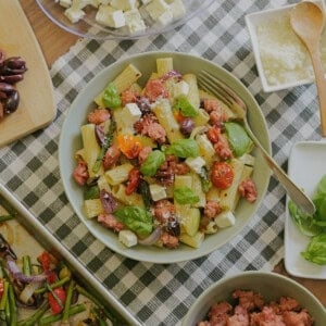 A bowl of rigatoni pasta with sausage, cherry tomatoes, olives, feta cheese, and basil sits on a checkered cloth, surrounded by ingredients like feta cubes, olives, sausage, veggies, and grated cheese.