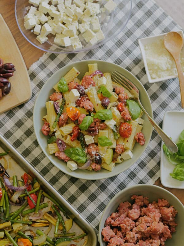 A bowl of rigatoni pasta with sausage, cherry tomatoes, olives, feta cheese, and basil sits on a checkered cloth, surrounded by ingredients like feta cubes, olives, sausage, veggies, and grated cheese.