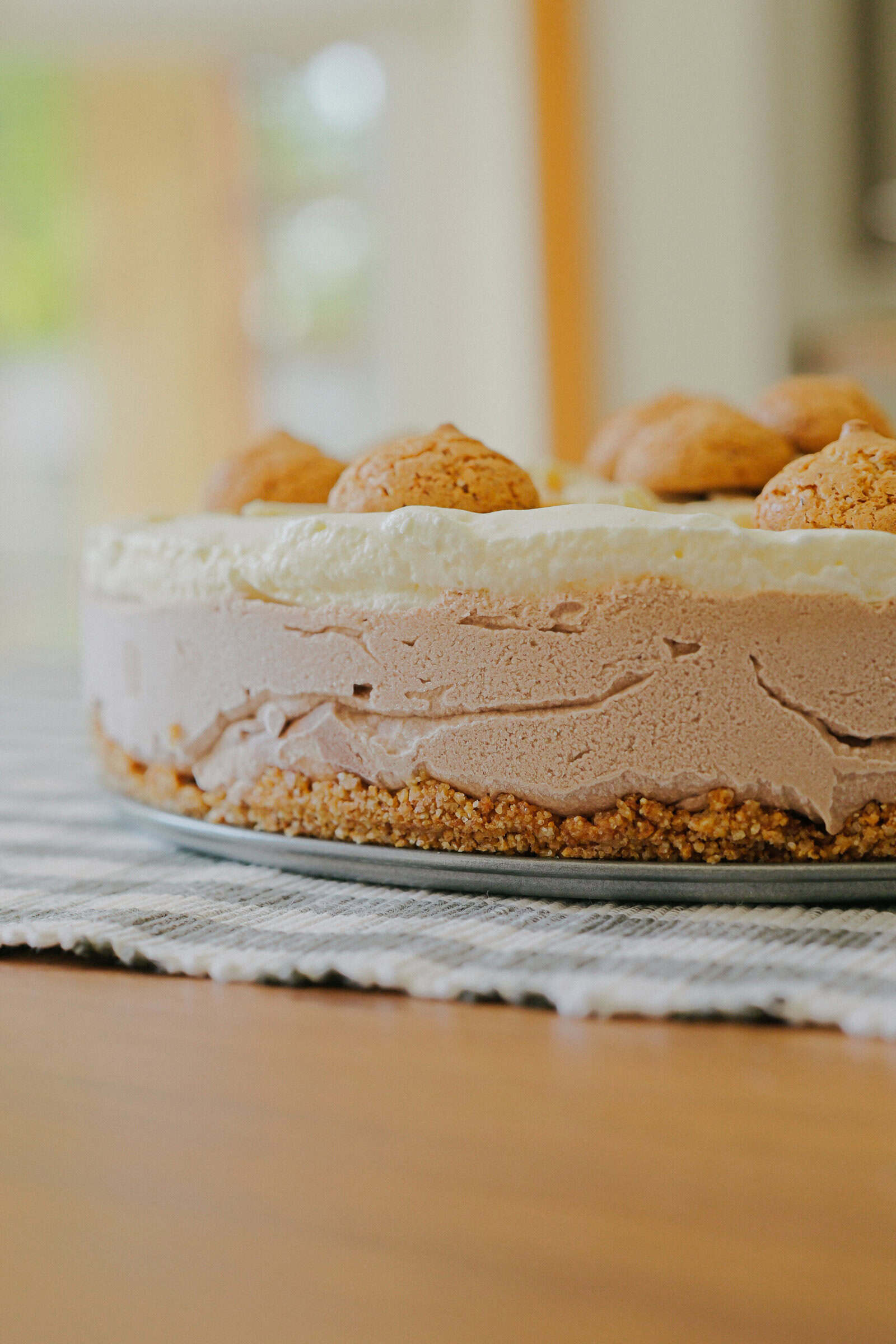 A close-up of a layered cheesecake with a crumbly crust, chocolate and vanilla layers, topped with round cookies, sitting on a striped cloth on a wooden table.