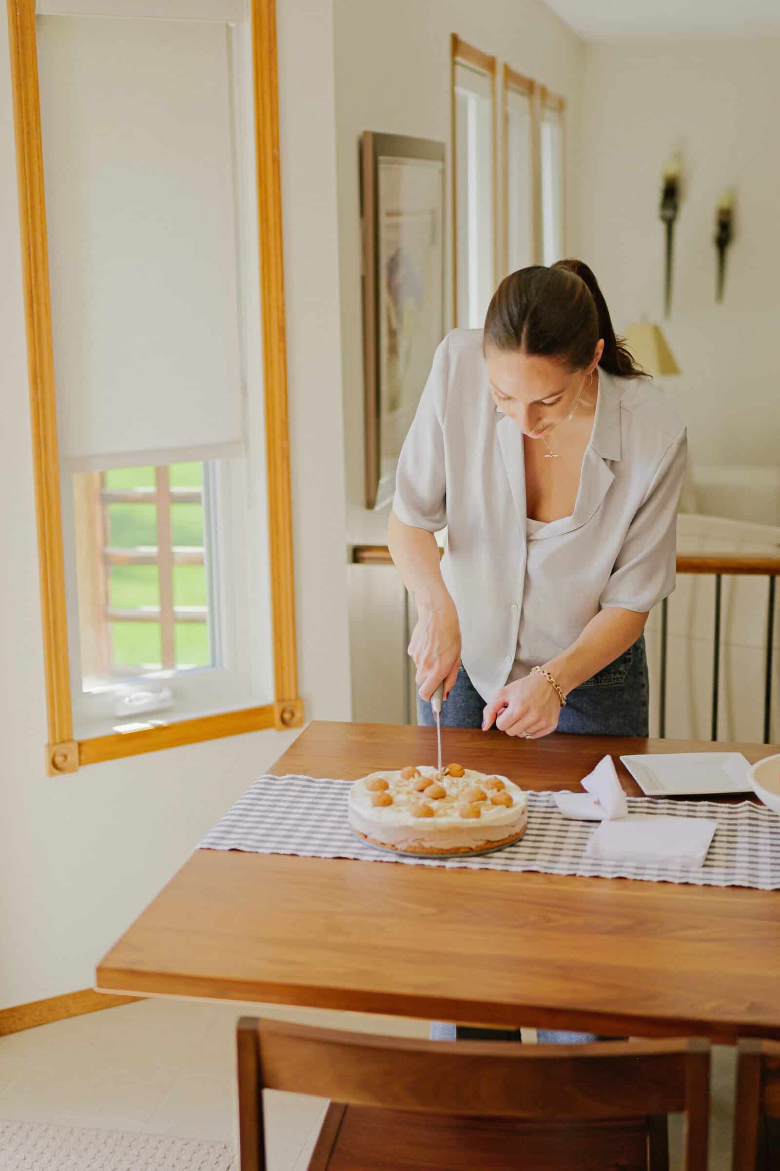 A woman stands at a wooden table in a bright room, cutting a cake topped with pastries. She is focused on slicing the cake, which sits on a checkered runner. Light streams in through the windows beside her.