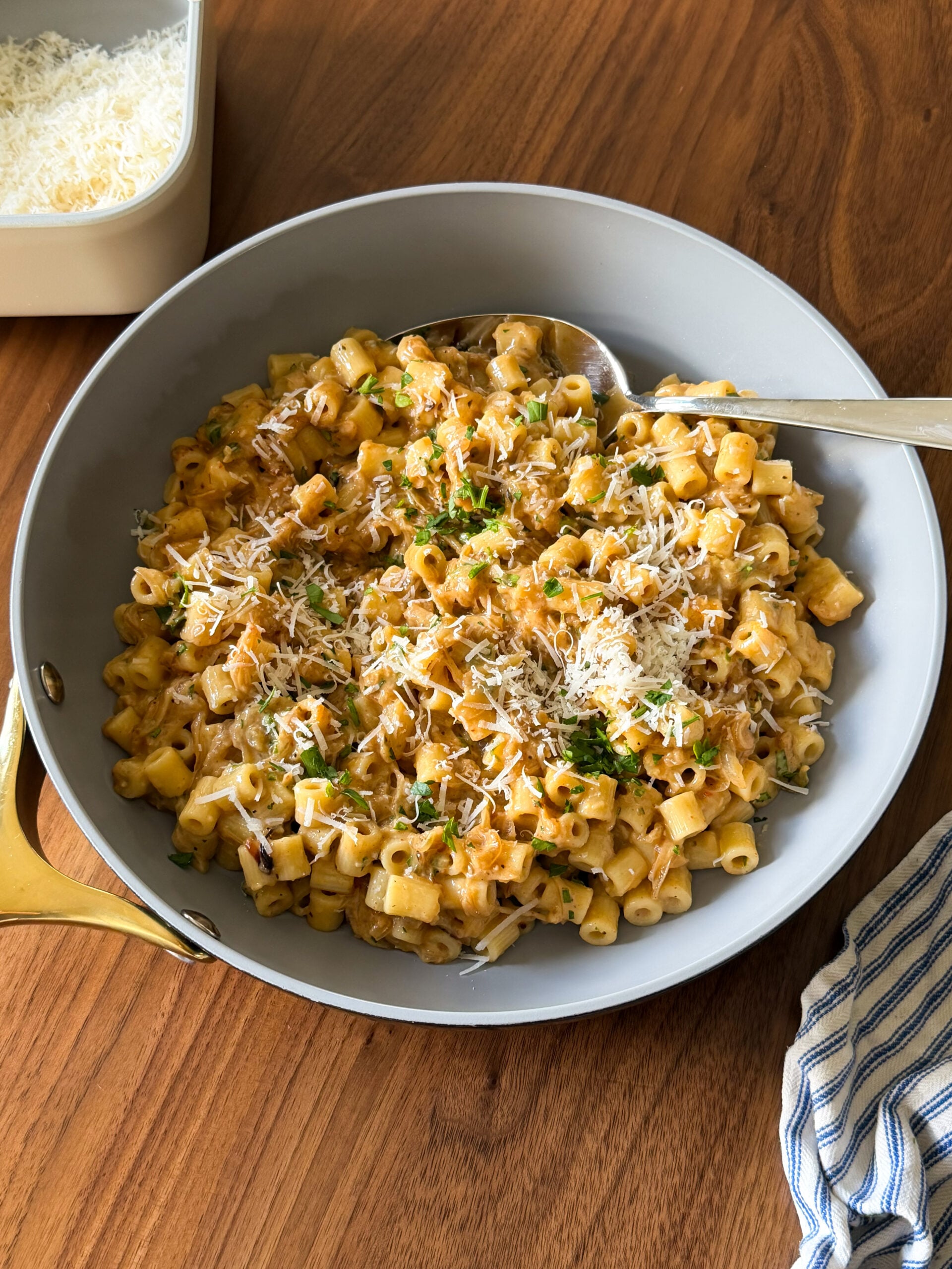 A skillet filled with creamy Caramelized Onion Pasta, topped with grated cheese and chopped herbs, sits on a wooden table, with a serving spoon and a striped cloth nearby. A dish of shredded cheese is also in view, making this One Pan Caramelized Onion Pasta recipe perfect for any cozy meal.