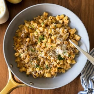 A pan of creamy pasta topped with grated cheese and herbs sits on a wooden table, next to a blue-striped cloth, onions, and a container of cheese—perfect for trying out a One Pan Caramelized Onion Pasta recipe.