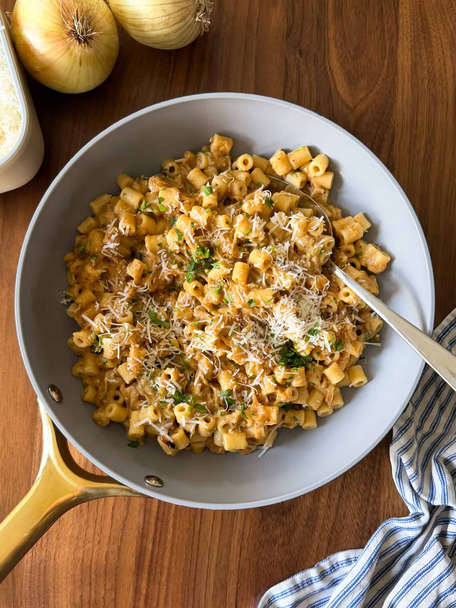A pan of creamy pasta topped with grated cheese and herbs sits on a wooden table, next to a blue-striped cloth, onions, and a container of cheese—perfect for trying out a One Pan Caramelized Onion Pasta recipe.