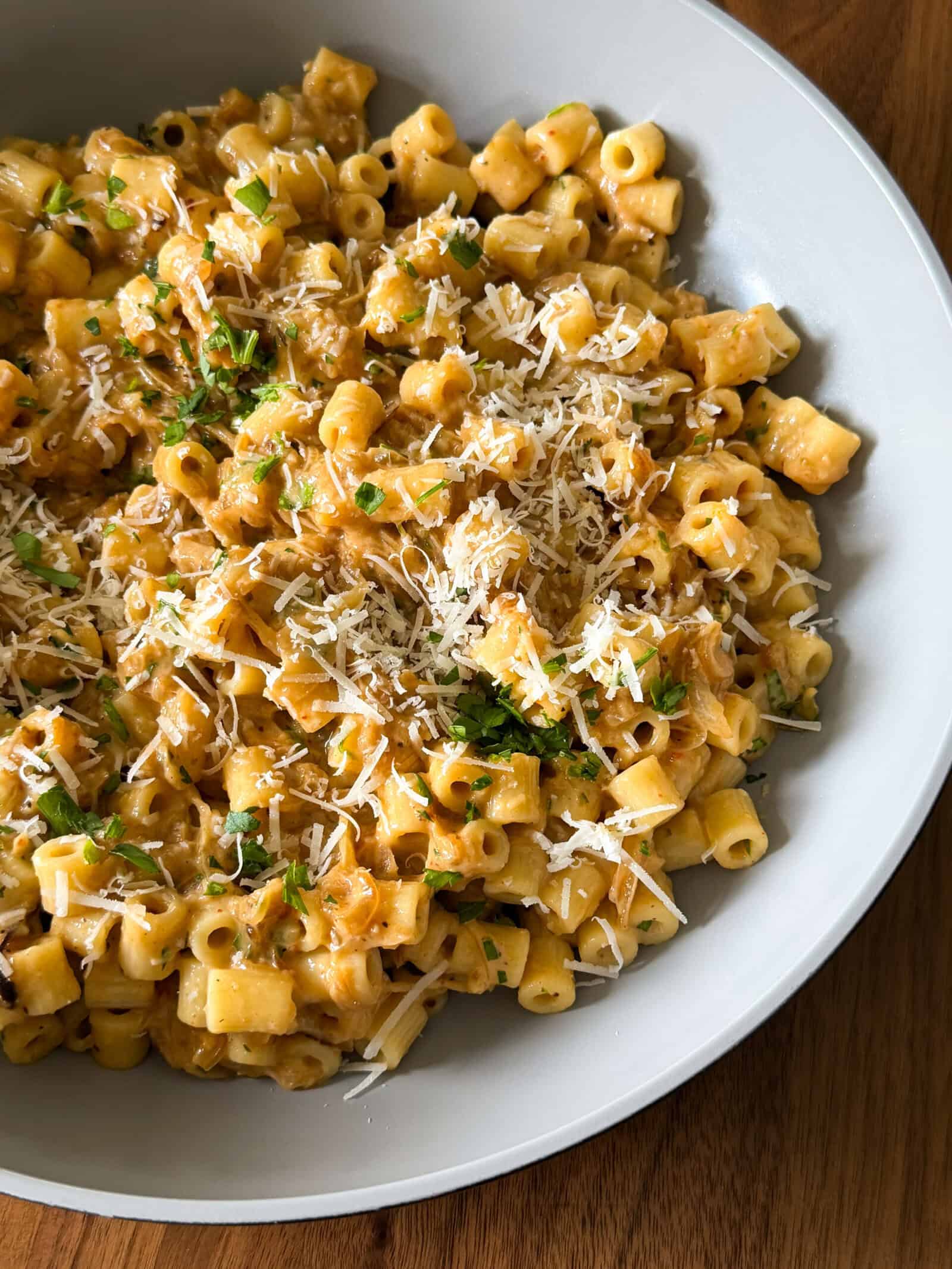 A bowl of creamy orange One Pan Caramelized Onion Pasta, topped with grated cheese and chopped herbs, sits on a wooden table.