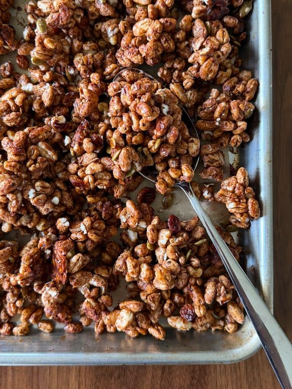 A baking tray filled with clusters of homemade granola made from puffed grains, nuts, and dried fruit, with a metal spoon resting in the mixture—a delicious example of a Homemade Cereal Recipe that’s both wholesome and easy to make.