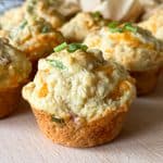Cheddar and chive savoury breakfast muffins rest on a wooden surface, their golden tops dotted with bits of green chive, surrounded by more breakfast muffins in the background.