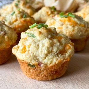 Cheddar and chive savoury breakfast muffins rest on a wooden surface, their golden tops dotted with bits of green chive, surrounded by more breakfast muffins in the background.