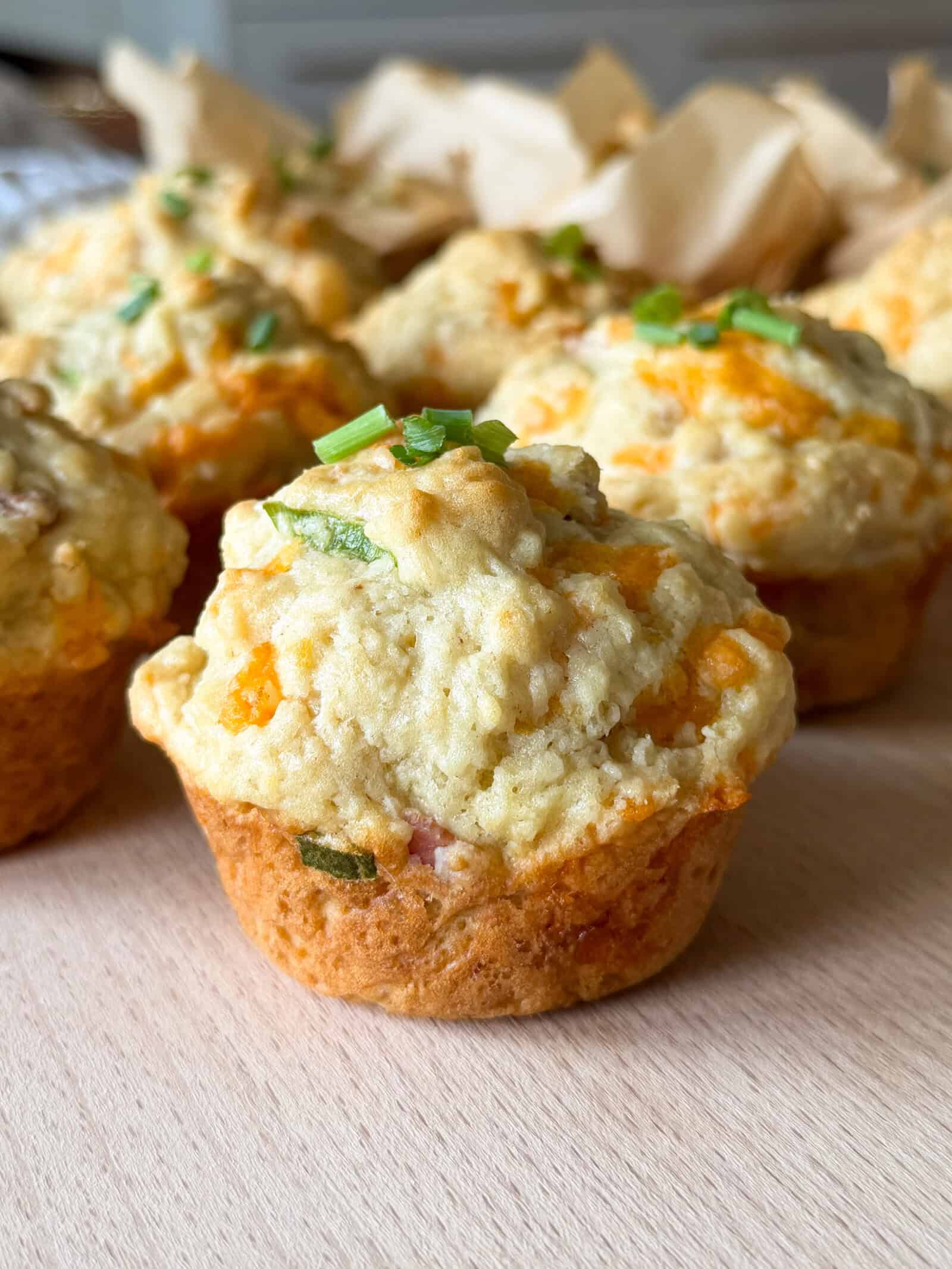 Cheddar and chive savoury breakfast muffins rest on a wooden surface, their golden tops dotted with bits of green chive, surrounded by more breakfast muffins in the background.