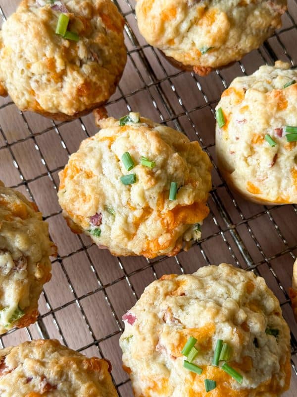 Cheddar and chive savoury breakfast muffins with bits of meat cooling on a wire rack, viewed from above. Some muffins are topped with chopped chives. The rack sits on a wooden surface—an inviting scene for anyone searching for the perfect breakfast muffins recipe.