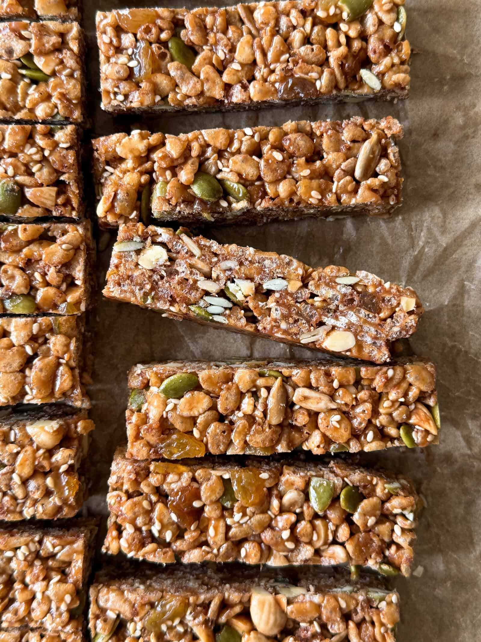 Several homemade breakfast bars, prepared with seeds and nuts, are arranged on parchment paper, with one bar placed on top of the others to showcase its textured surface. This delicious breakfast bars recipe makes for a wholesome and satisfying start to your morning.