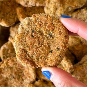 A hand with blue fingernails holds a round, golden-brown breaded nugget in front of a pile of similar nuggets, freshly prepared using a Homemade Chicken Nuggets Recipe.
