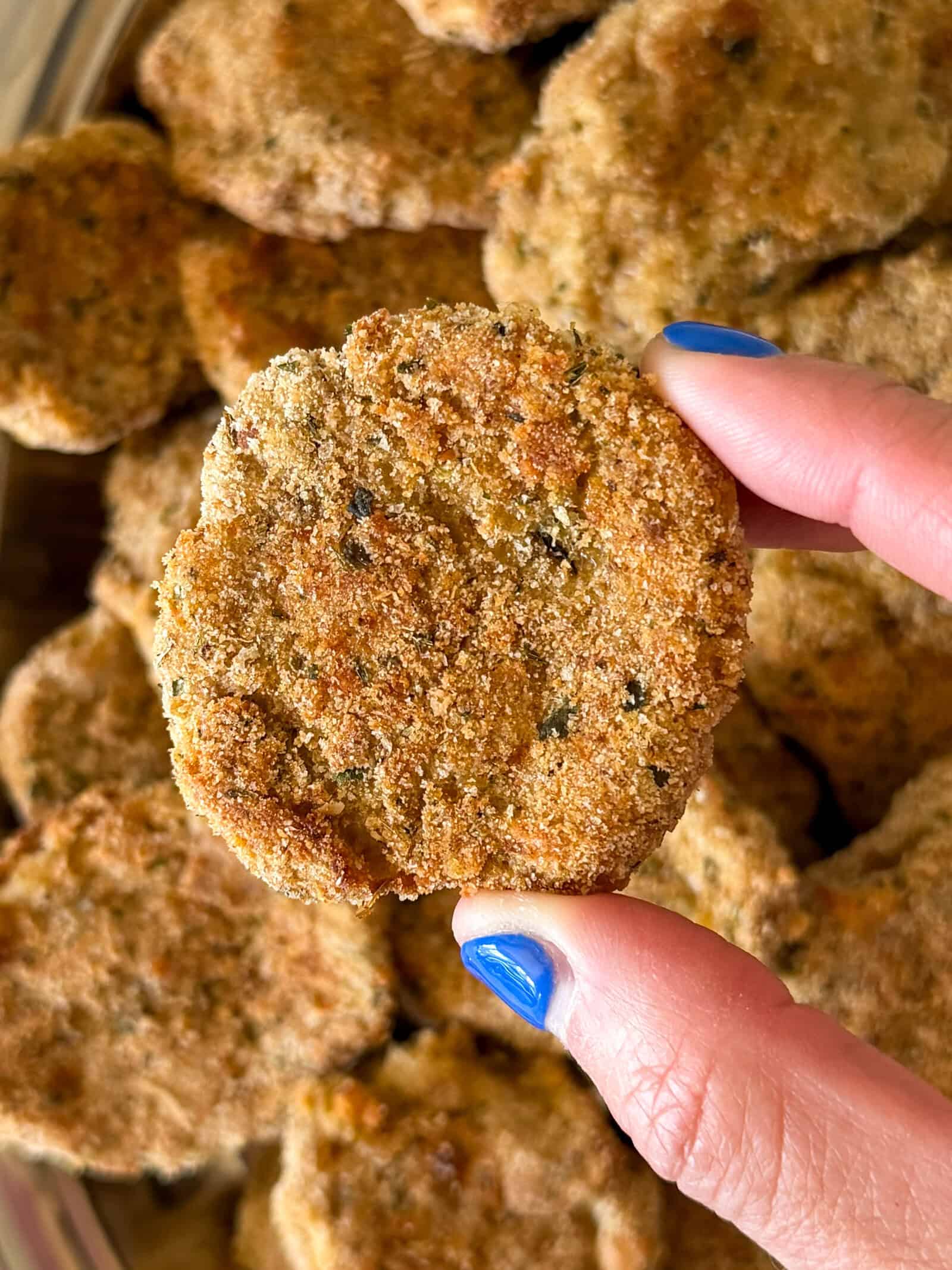 A hand with blue fingernails holds a round, golden-brown breaded nugget in front of a pile of similar nuggets, freshly prepared using a Homemade Chicken Nuggets Recipe.