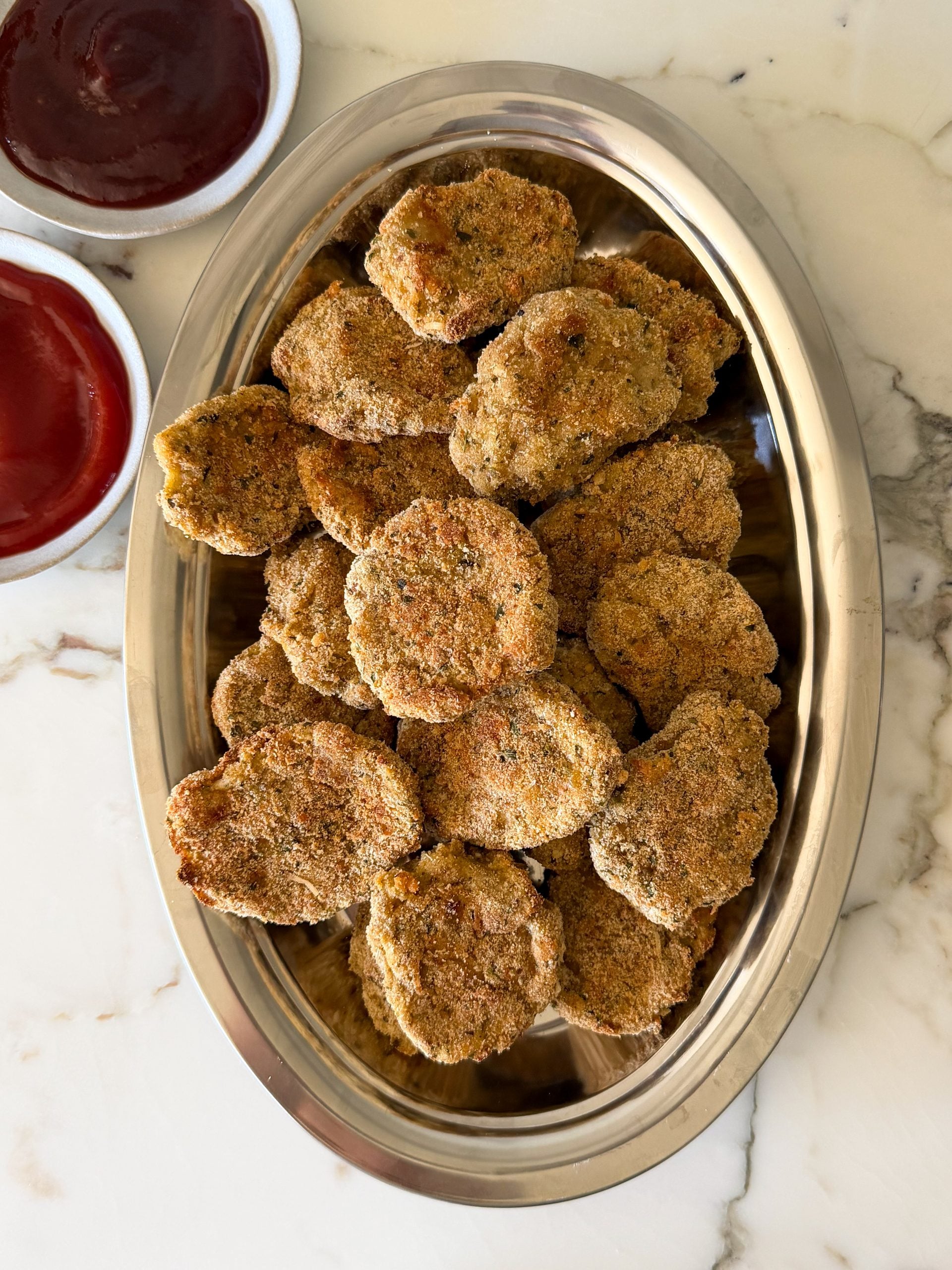 A silver tray filled with breaded, baked or fried vegetable patties, reminiscent of a Homemade Chicken Nuggets Recipe, with two bowls of dipping sauces—one red, one dark red—on a marble surface.