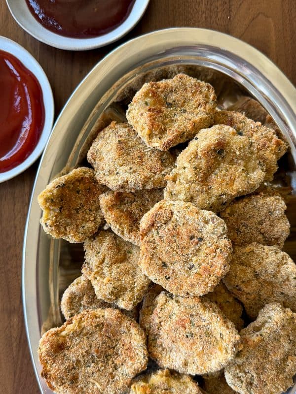 A silver tray filled with breaded, golden-brown homemade chicken nuggets sits on a wooden table, with two small dishes of red dipping sauce beside it.