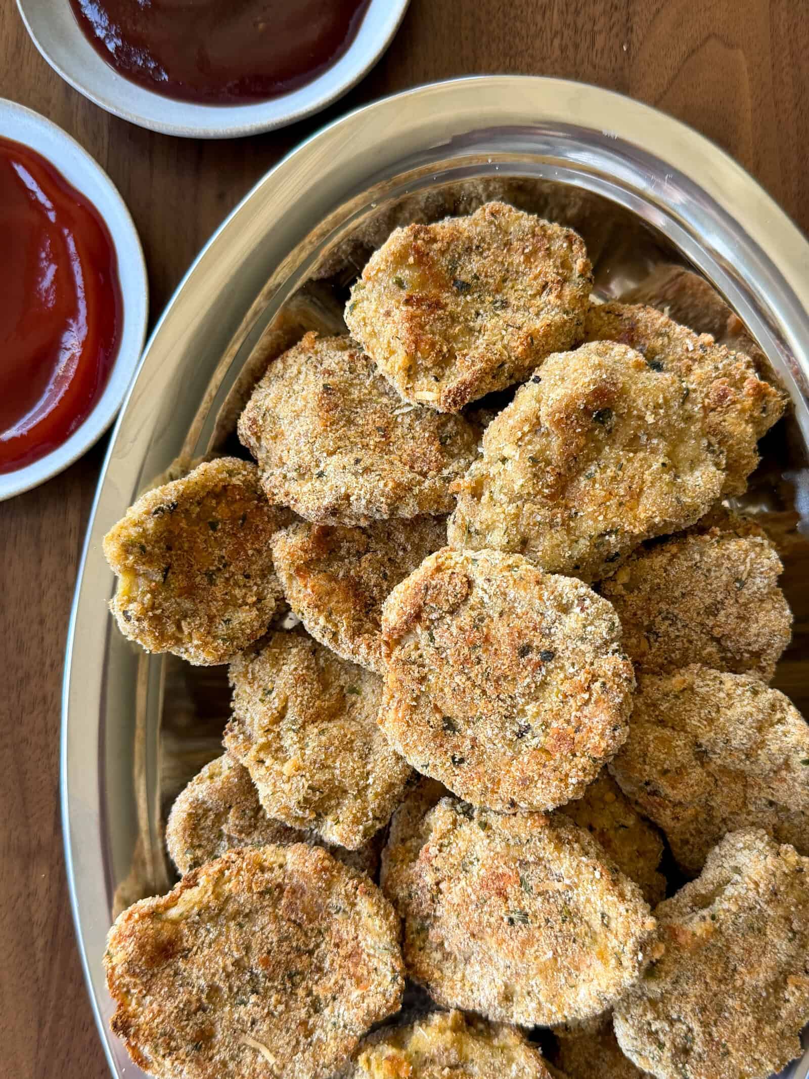A silver tray filled with breaded, golden-brown homemade chicken nuggets sits on a wooden table, with two small dishes of red dipping sauce beside it.
