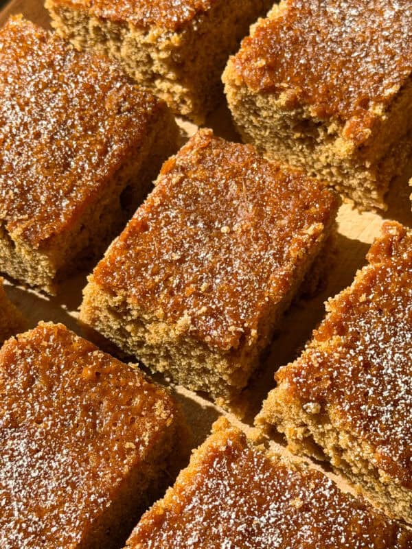 Nine slices of moist, golden-brown Gingerbread Cake topped with powdered sugar are arranged closely together on a wooden surface, highlighted by warm sunlight—a tempting glimpse of a classic gingerbread cake recipe brought to life.