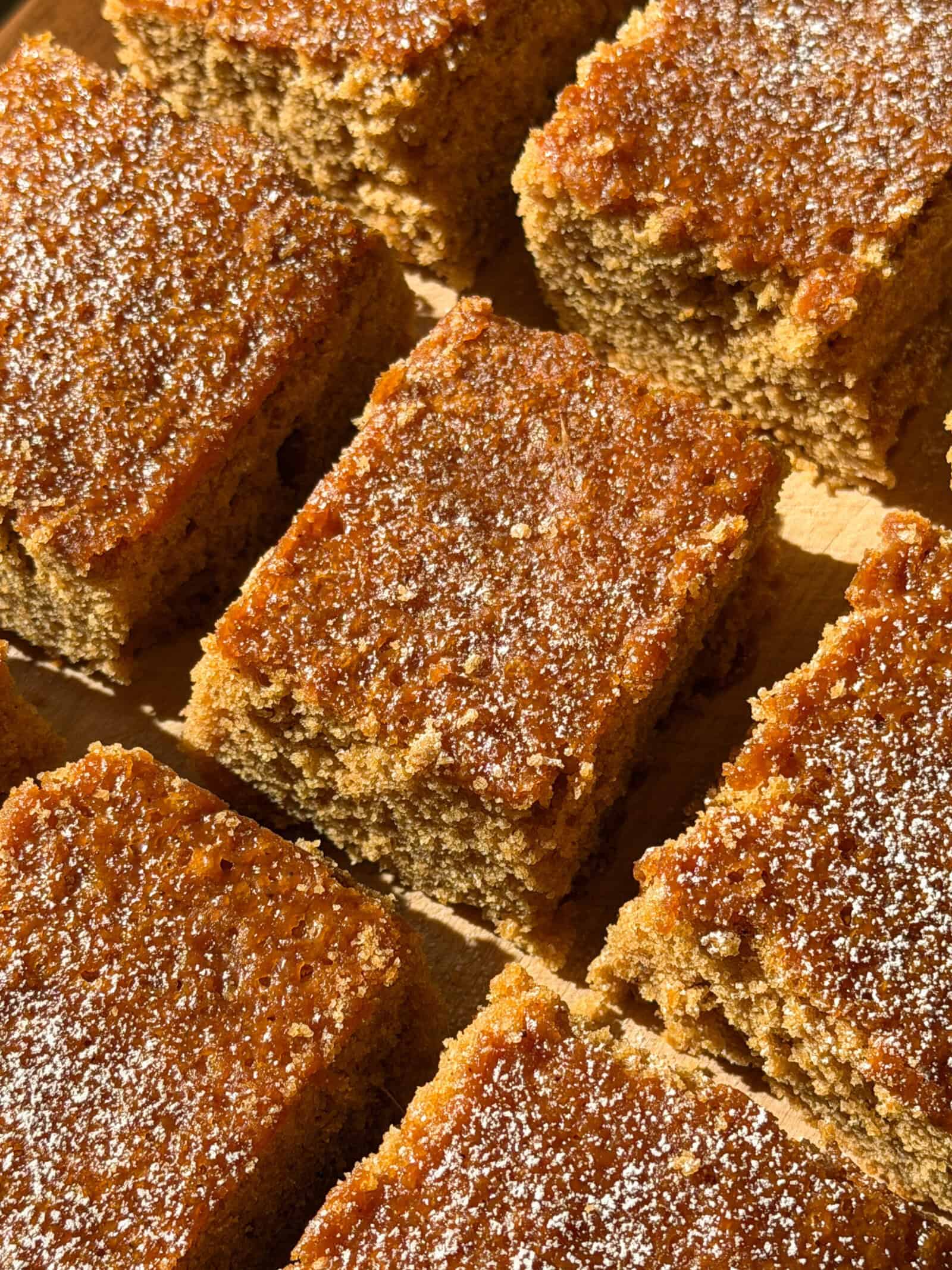 Nine slices of moist, golden-brown Gingerbread Cake topped with powdered sugar are arranged closely together on a wooden surface, highlighted by warm sunlight—a tempting glimpse of a classic gingerbread cake recipe brought to life.