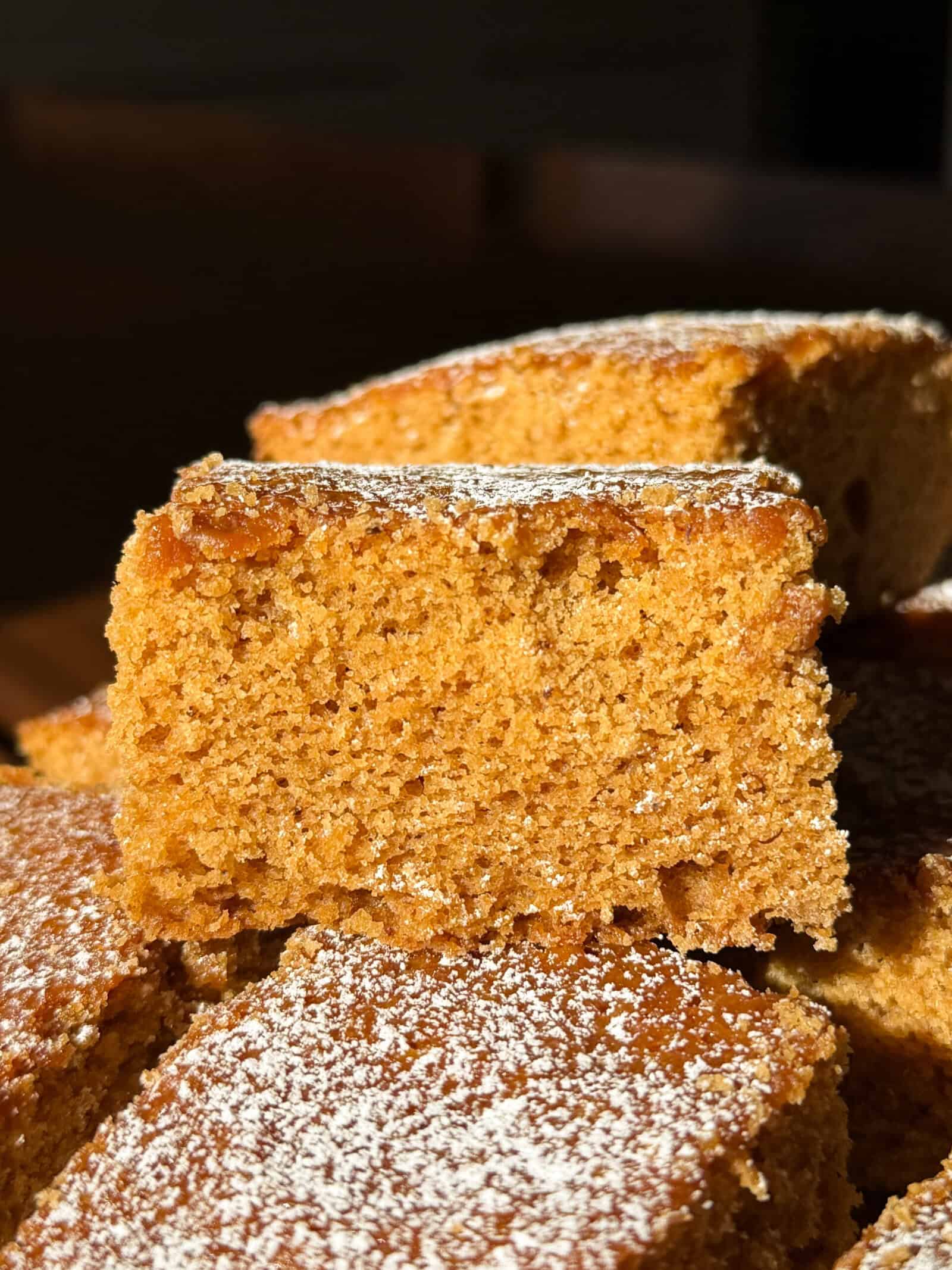 A close-up of golden brown Gingerbread Cake slices stacked, dusted with powdered sugar, with soft, moist texture visible in bright sunlight.
