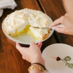 A person cuts a slice from an Easy Lemon Meringue Pie, holding the pie tin over a wooden table with an empty plate nearby.