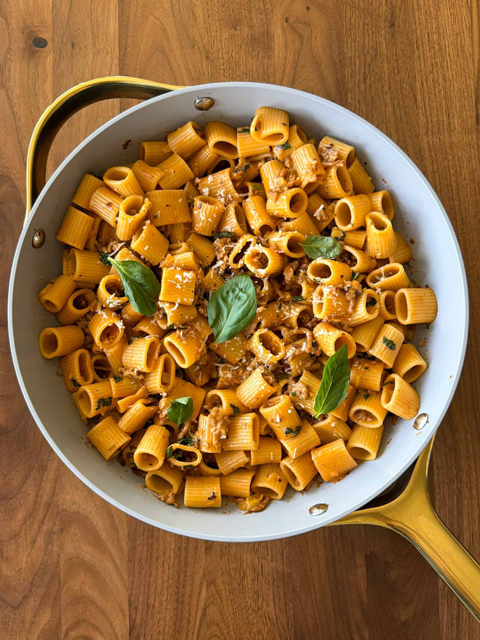 A skillet filled with rigatoni pasta in an orange sauce, garnished with fresh basil leaves and crumbled cheese, sits on a wooden surface—an inviting scene that perfectly captures the essence of a comforting one pot pasta, such as One Pot Sausage and Fennel Pasta.