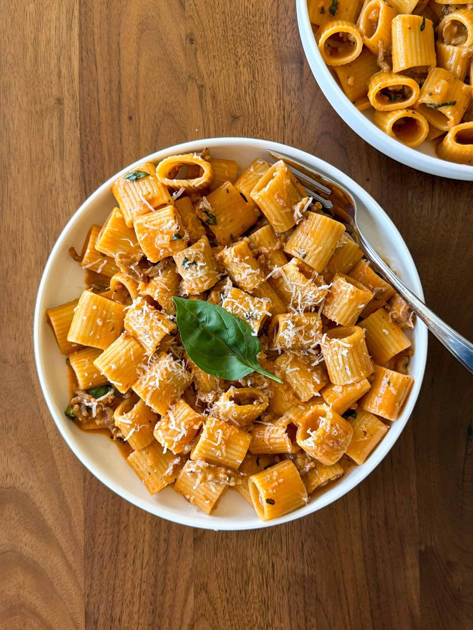 A bowl of rigatoni pasta with tomato sauce, grated cheese, and fresh basil on top sits on a wooden table with a fork and part of another pasta bowl visible, reminiscent of a cozy one pot pasta meal.