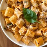 A bowl of rigatoni pasta in a creamy tomato sauce, inspired by a One Pot Sausage and Fennel Pasta, topped with grated parmesan cheese and a fresh basil leaf, served on a wooden table.