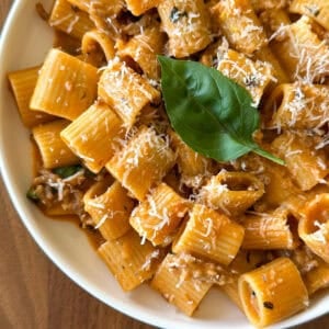 A bowl of rigatoni pasta in a creamy tomato sauce, inspired by a One Pot Sausage and Fennel Pasta, topped with grated parmesan cheese and a fresh basil leaf, served on a wooden table.