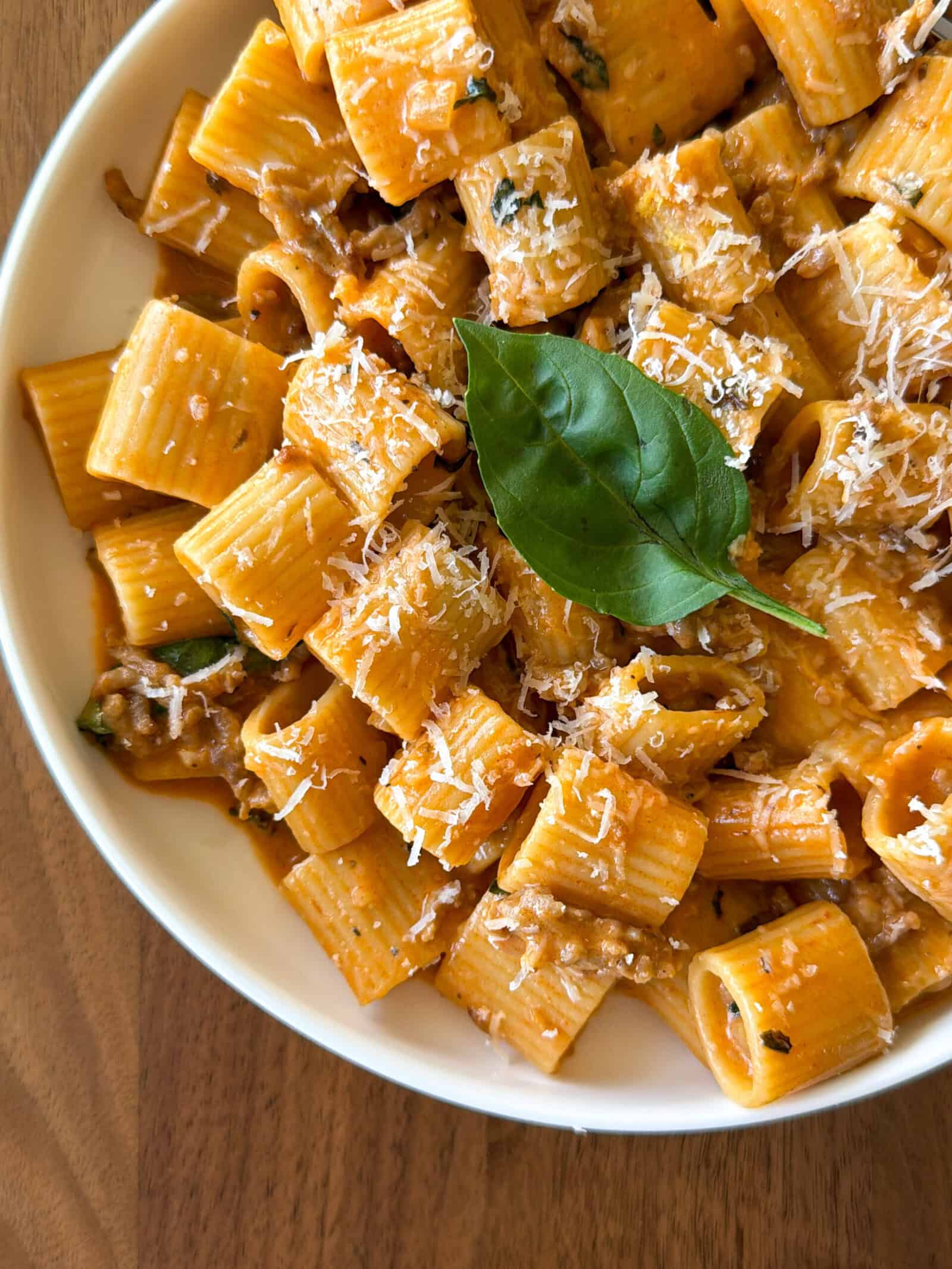 A bowl of rigatoni pasta in a creamy tomato sauce, inspired by a One Pot Sausage and Fennel Pasta, topped with grated parmesan cheese and a fresh basil leaf, served on a wooden table.