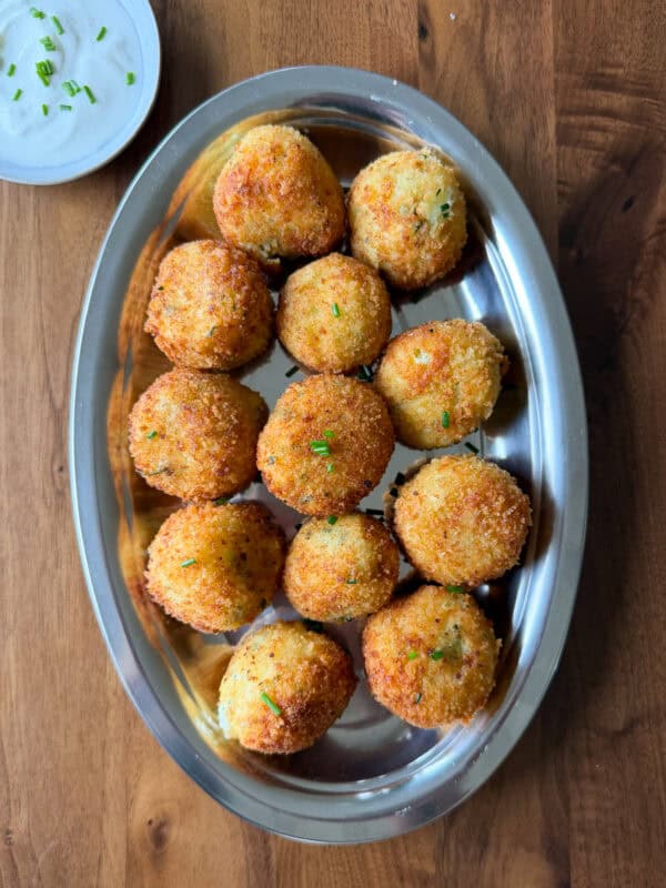 A metal tray with 13 golden-brown, round mashed potato balls garnished with green herbs. A small dish of white dipping sauce sits nearby on a wooden table—an irresistible solution for anyone wondering what to do with leftover mashed potatoes.