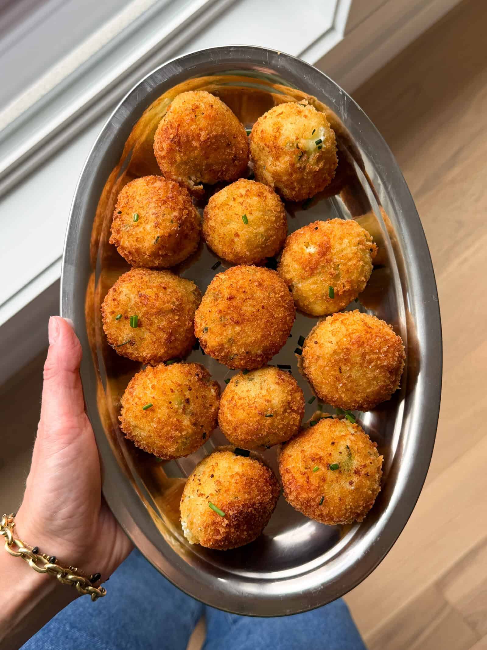 A hand holds a silver oval tray filled with twelve golden-brown, crispy fried mashed potato balls, garnished with chopped herbs, above a wooden floor&mdash;a delicious idea for what to do with leftover mashed potatoes.
