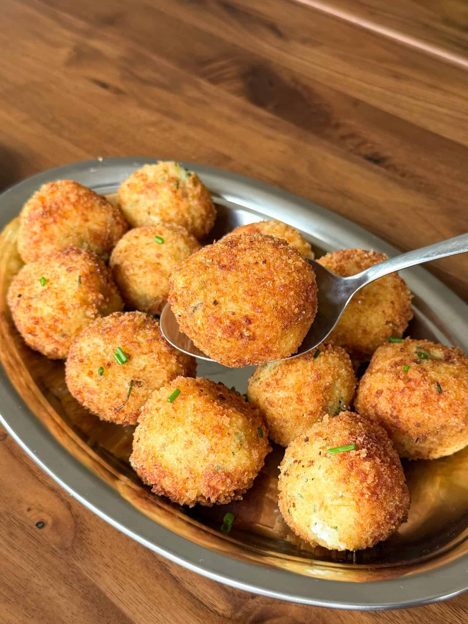 A metal platter with several golden-brown, crispy fried mashed potato balls. A spoon holds up one ball, showing its round shape and crunchy coating. The balls are garnished with small green herb pieces&mdash;an irresistible idea for what to do with leftover mashed potatoes.