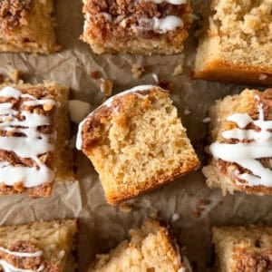 Nine square pieces of easy coffee cake with crumb topping and white icing drizzle are arranged on brown parchment paper, with one piece turned to show its moist inside texture—a perfect example of a classic cinnamon coffee cake recipe.
