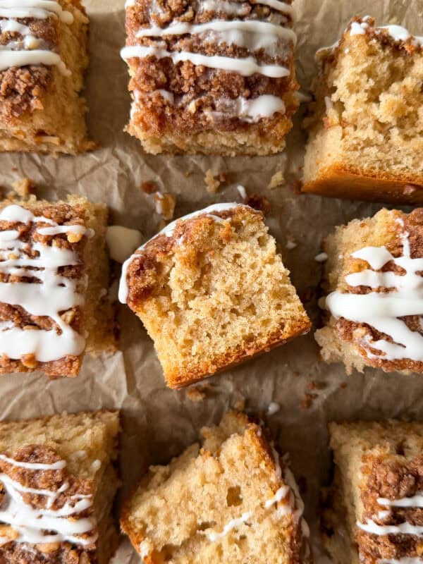 Nine square pieces of easy coffee cake with crumb topping and white icing drizzle are arranged on brown parchment paper, with one piece turned to show its moist inside texture—a perfect example of a classic cinnamon coffee cake recipe.