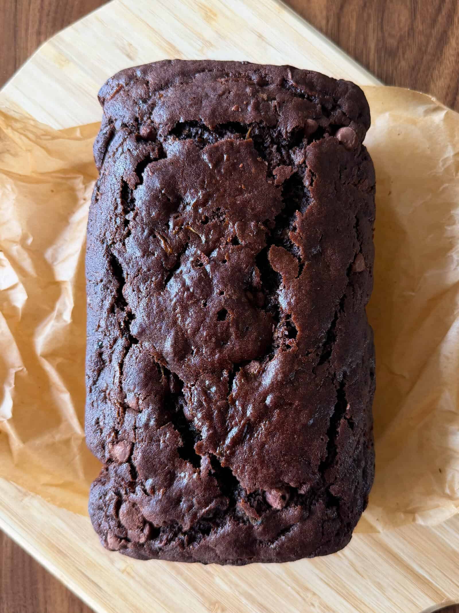 A loaf of rich, dark Chocolate Zucchini Bread rests on brown parchment paper atop a wooden cutting board. The bread has a cracked, textured top and appears moist and dense—an irresistible result from our favorite Chocolate Zucchini Bread Recipe.