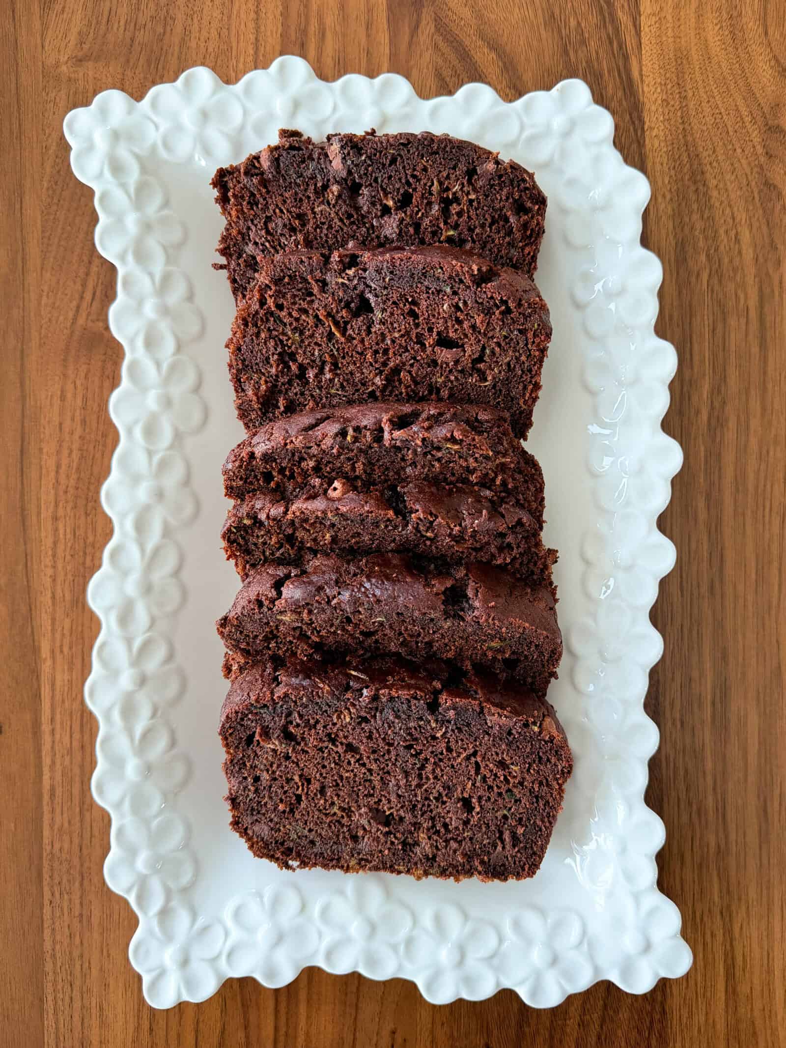 Five thick slices of Chocolate Zucchini Bread are arranged on a white, flower-edged rectangular plate, set on a wooden surface. This irresistible treat is perfect for anyone searching for a comforting Chocolate Zucchini Bread Recipe.
