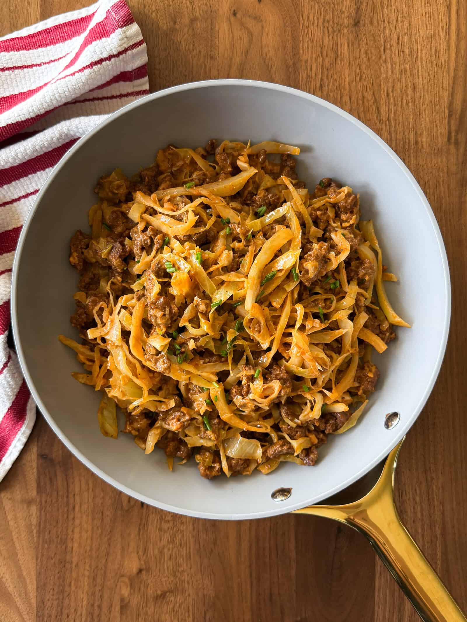 A skillet on a wooden table contains cooked ground meat, onions, and noodles, garnished with herbs in a delicious homemade hamburger helper recipe. A red and white striped towel is next to the skillet.