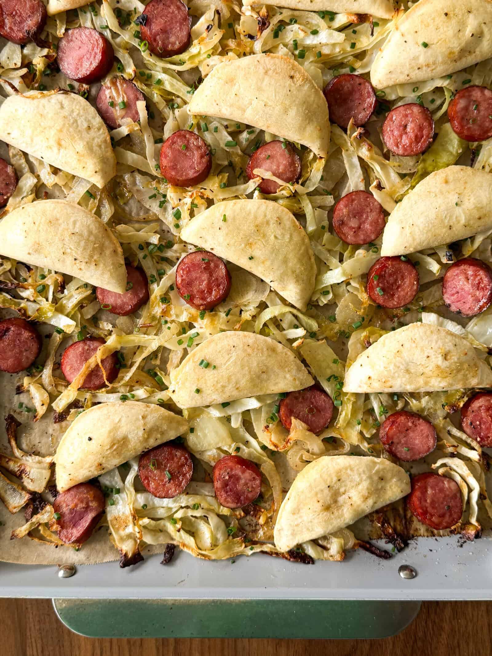 Sheet pan pierogis and kielbasa featuring roasted cabbage, sliced sausage rounds, and folded flatbreads topped with chopped chives.