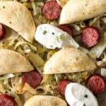 Sheet pan pierogis and kielbasa featuring sliced sausage and cabbage topped with folded tortillas, sour cream dollops, and chives.