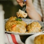 A hand with blue nail polish holds a fork with a bite of creamy, baked casserole above a patterned plate filled with similar portions.