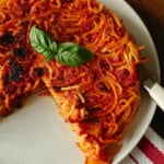 A round spaghetti pie with tomato sauce and basil garnish, served on a white plate. A slice has been cut and removed, revealing the pies inside. The plate sits on a red and white striped cloth.