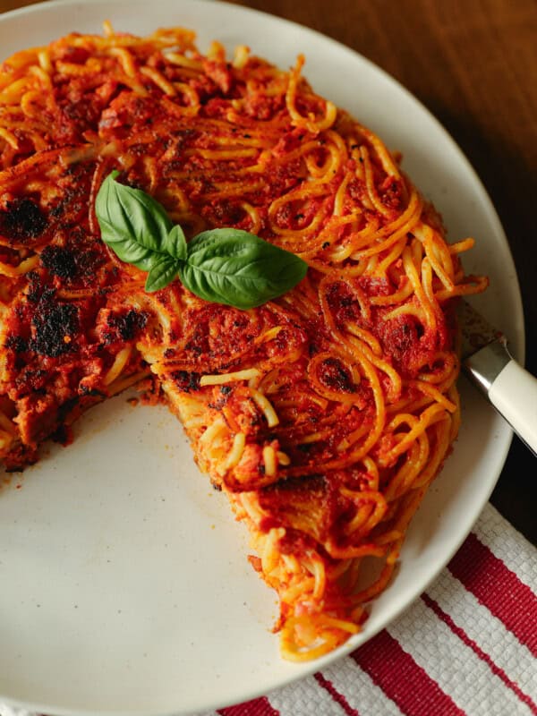 A round spaghetti pie with tomato sauce and basil garnish, served on a white plate. A slice has been cut and removed, revealing the pies inside. The plate sits on a red and white striped cloth.