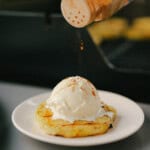 A hand sprinkles cinnamon onto a scoop of vanilla ice cream placed on a grilled pineapple slice, served on a small white plate.