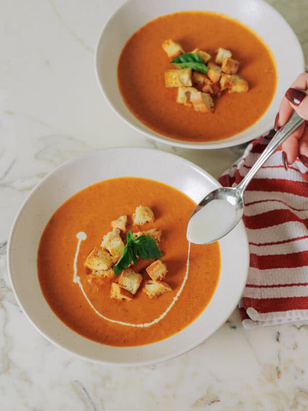 A hand uses a spoon to drizzle cream into a bowl of roasted tomato soup, served alongside another bowl topped with croutons and basil.
