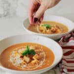 A hand sprinkles grated cheese over a bowl of roasted tomato soup topped with croutons and a basil leaf, with a second bowl of soup nearby.