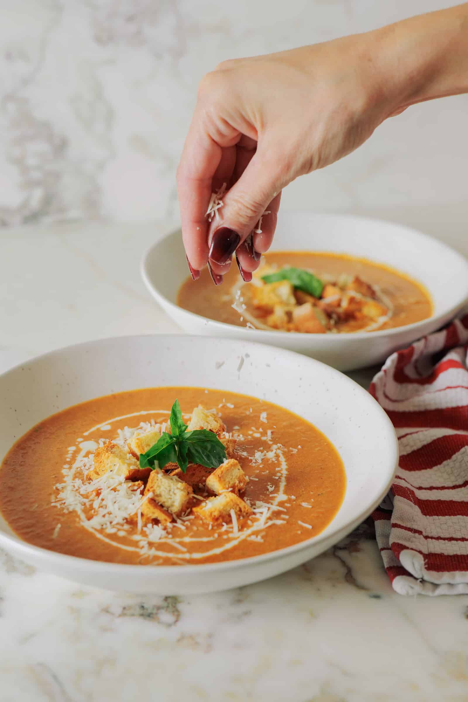 A hand sprinkles grated cheese over a bowl of roasted tomato soup topped with croutons and a basil leaf, with a second bowl of soup nearby.