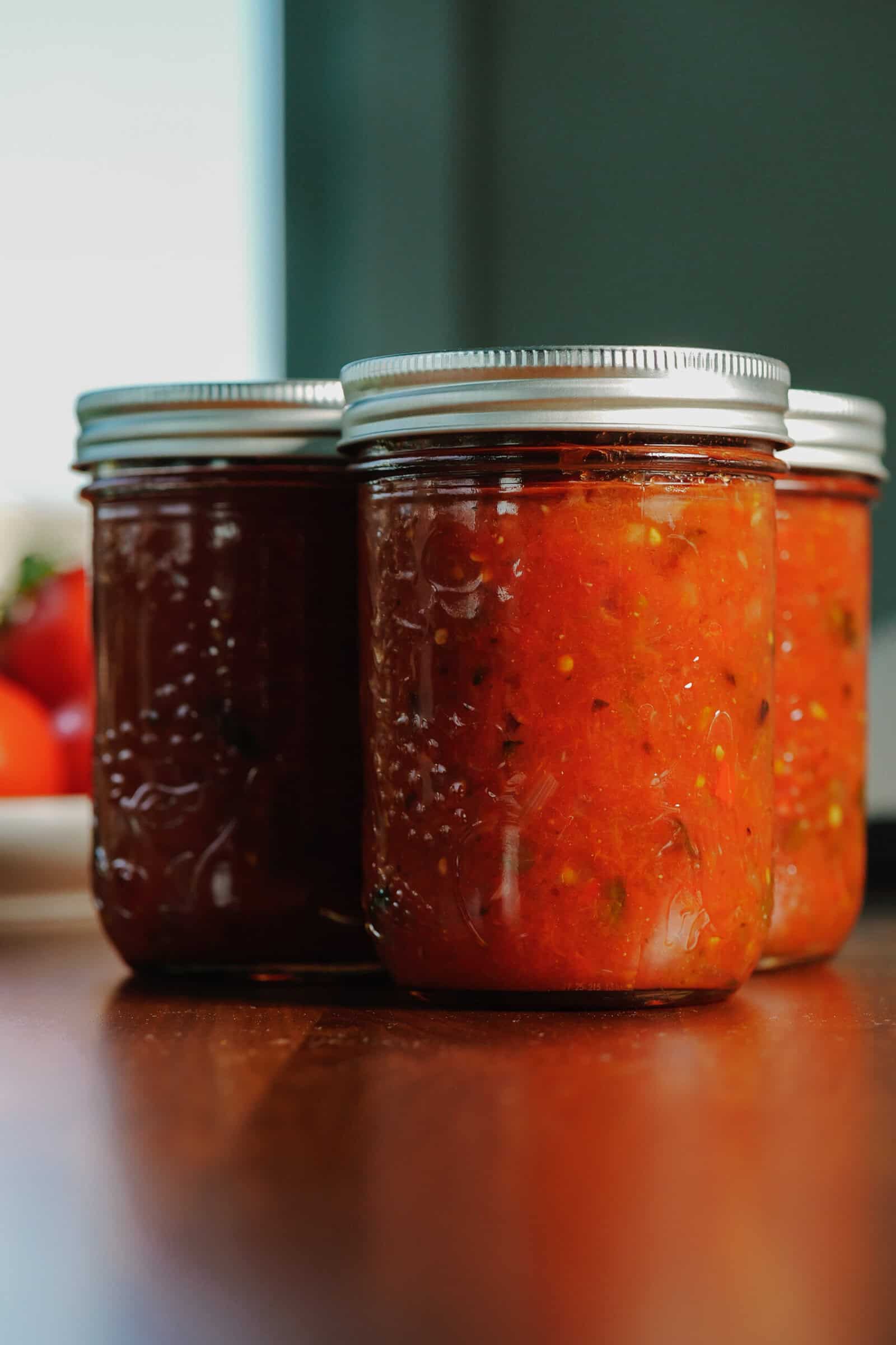 Three glass jars of homemade tomato sauce with silver lids sit on a wooden surface, evoking the comforting feeling of a classic canned salsa recipe, with blurred tomatoes visible in the background.